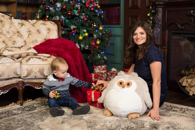 Mother and son open Christmas gifts on the carpet near the Christmas tree and the fireplace