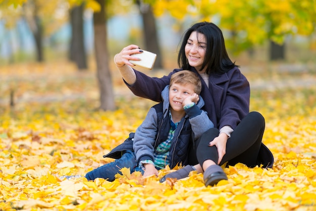 Mother and son posing for a selfie in a park sitting amongst colorful yellow autumn leaves smiling for the camera