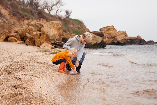 Mother and son sitting on the beach playing with paper boat in autumn time