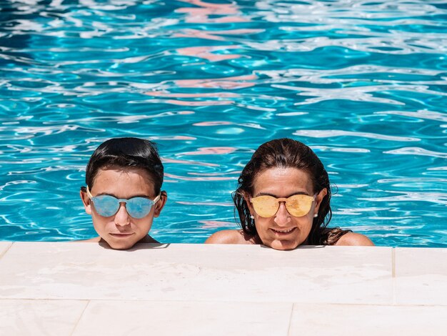 Mother and son with sunglasses are leaning on the edge of the pool on a sunny summer day