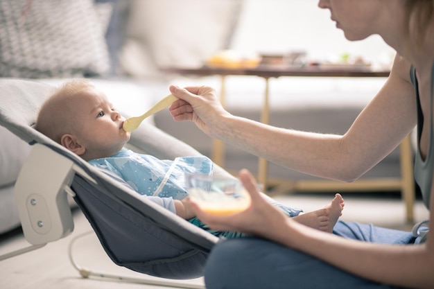 Mother spoon feeding her baby boy child in baby chair with fruit puree baby solid food introduction