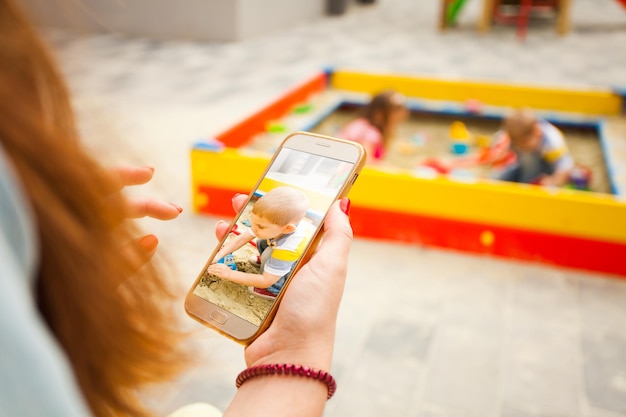 Mother taking photo of her son at the smartphone. Children play at sandbox on the background, posting in social media