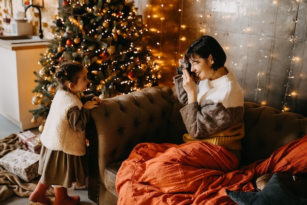 Mother taking pictures of her daughter at home, next to a Christmas tree