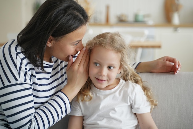 Mother telling secret whispering to kid daughter gossiping together at home Trustful conversation