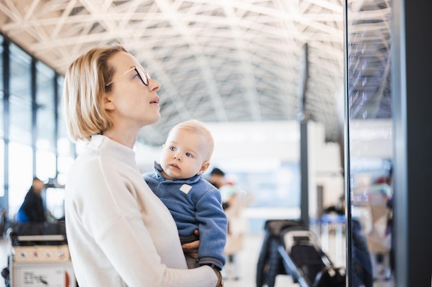 Mother traveling with child holding his infant baby boy at airport terminal checking flight schedule waiting to board a plane Travel with kids concept