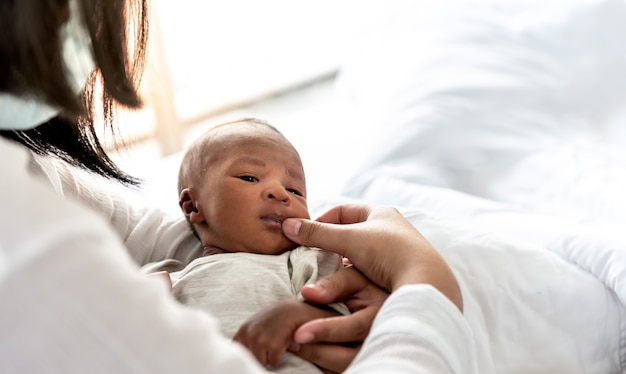 Mother wipes the milk stain with her fingers on the corner of the son's mouth, is African baby newborn 12 day old, in white bed in the bedroom, to African family and black skin newborn concept.