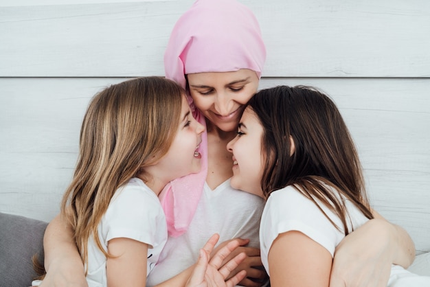 A mother with cancer wears a pink headscarf, tenderly and happily hugging her two blond and brown-haired daughters.