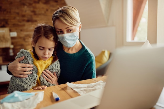 Mother with face mask using laptop while taking care of her ill daughter at home