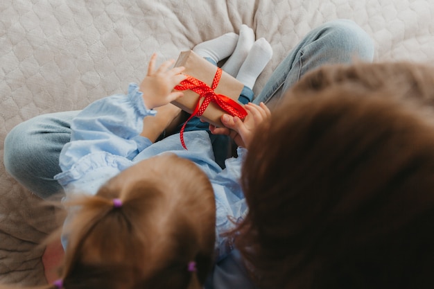 Mother with her cute little daughter sitting on the bed, holding a gift box in their hands. Top view. Close-up.