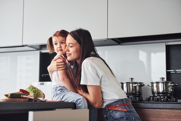 Mother with her little daughter embracing each other indoors in kitchen