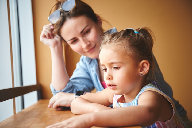 Mother with her little daughter sitting near window in to the cafe. Waiting for the order