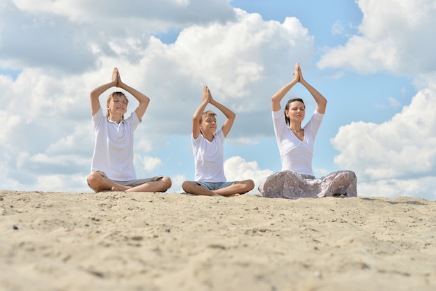 Mother with her sons sitting on a sand and doing yoga