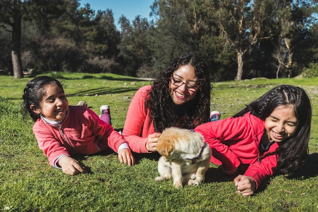 Mother with her two daughters lying on the grass and playing with golden retriever puppy in the park