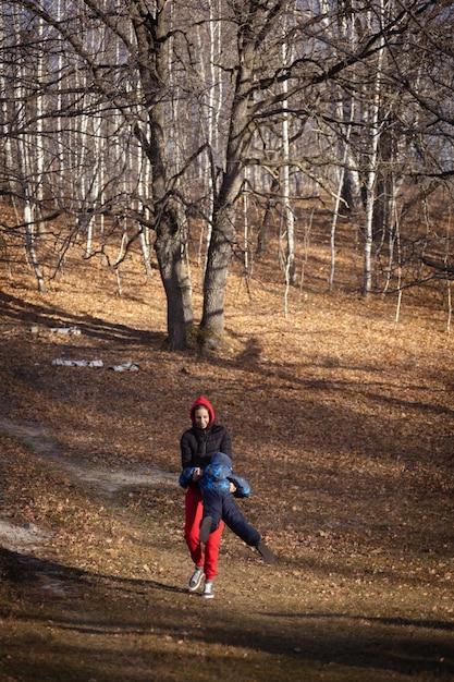 Mother with little son having fun while on walk on forest country road with dry leaves fallen on ground from trees in background on sunny autumn day Family time together