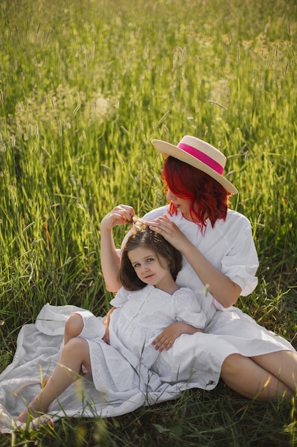 Mother with red hair and a hat is sitting in a green field with grass with her daughter of four years at sunset