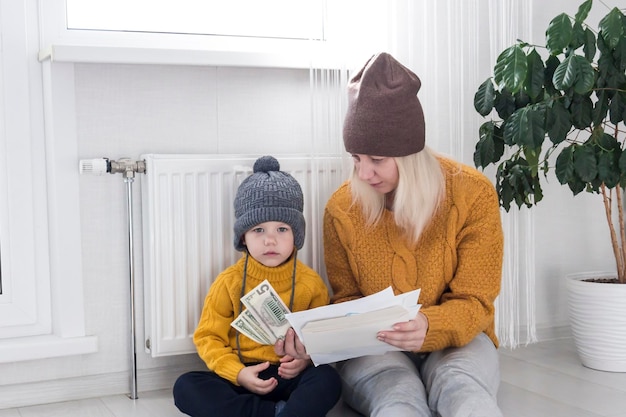 A mother with a small child in a yellow sweater and hats is counting money and thinking how to pay bills and taxes near a heater with a thermostat