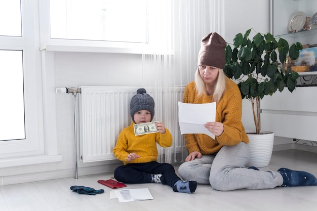 A mother with a small child in a yellow sweater and hats is counting money and thinking how to pay bills and taxes near a heater with a thermostat