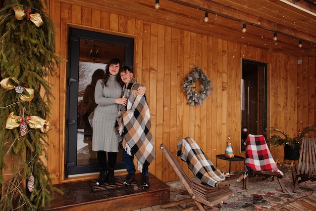 Mother with son in covered blanket in winter day against wooden house