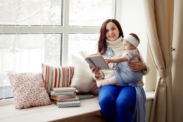 Mother and wrapped baby girl sit on window sill and read books at cozy winter, happy family leisure