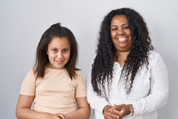 Mother and young daughter standing over white background with hands together and crossed fingers smiling relaxed and cheerful. success and optimistic