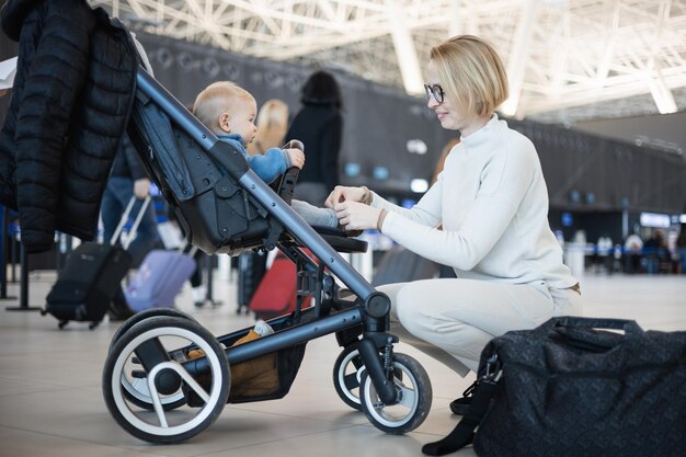 Motherat interacting with her infant baby boy child in stroller while travelling at airport terminal station Travel with child concept