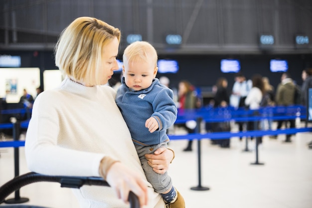 Motherat travelling with his infant baby boy child walking pushing baby stroller and luggage cart at airport terminal station Travel with child concept