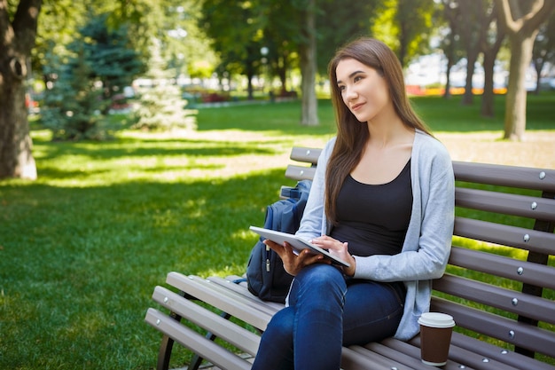 Motivated asian freelancer in the park. Young brunette girl on a bench, drinking coffee and working with tablet online. Lifestyle, technology and remote work concept