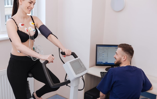 Motivated charming young lady feeling great while doing some exercises and the doctor examining her performance results