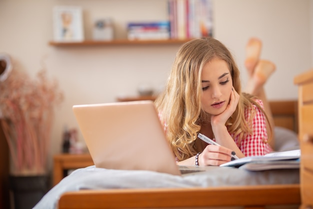 Photo motivated teen student lying on a bed solving assignements and having an online class from home due to global coronavirus pandemic.