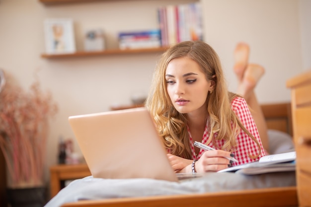 Motivated teen student lying on a bed solving assignements and having an online class from home due to global coronavirus pandemic.