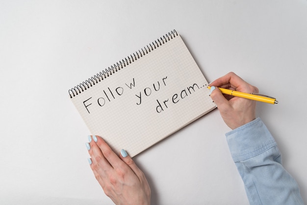 Motivational inscription in notebook Follow your dream. Female hands writing in notepad. White .