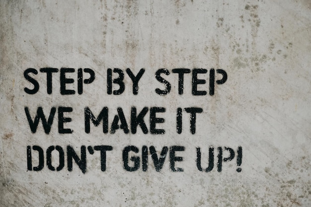 Motivational quote "step by step we make it, don't give up" written on a white wall with black paint