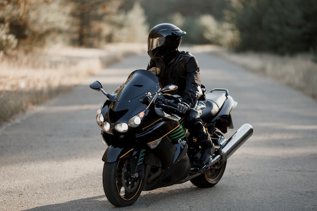 Motorcycle driver in a leather suit and sitting on a motorcycle alone on an asphalt highway against a forest background