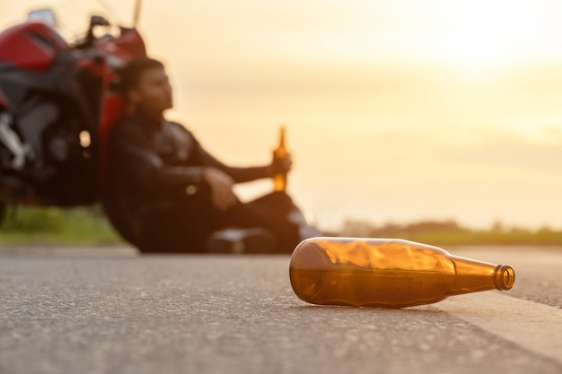 Motorcyclist sitting on the road beside his motorcycle, drinking an alcohol or beer