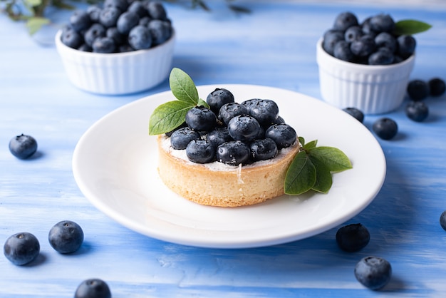 Mouth-watering blueberry tart in a white plate on a blue background, delicious berry dessert.