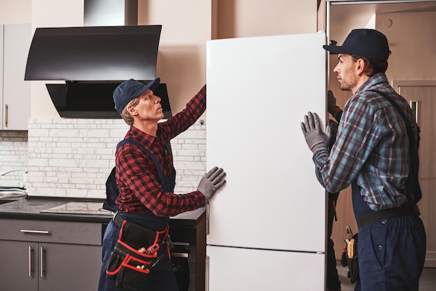 Moving again. men mechanics moving refrigerator to another place. They assist each other. Side view