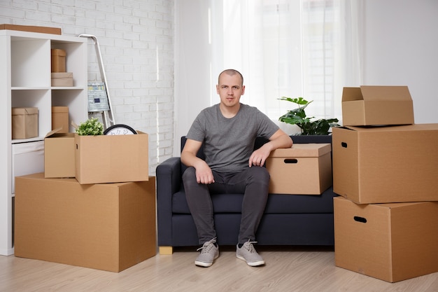 Moving concept - happy man sitting on sofa in room after moving day