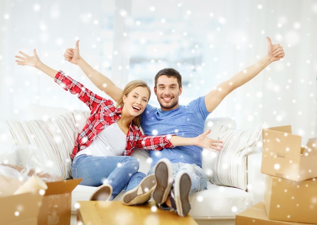 moving, home and couple concept - smiling couple showing thumbs up and waving hands on sofa in new home