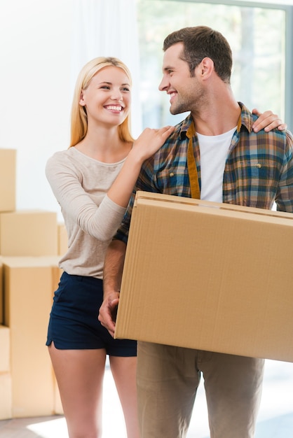 Moving to a new house together. Happy young man holding a cardboard box while his girlfriend holding hands on his shoulders