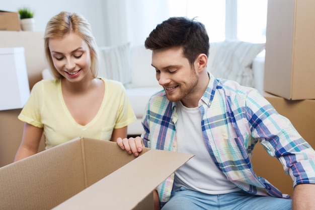 Photo moving, people, repair and real estate concept - smiling couple looking into big cardboard box at new home