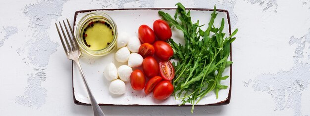 Mozzarella, cherry tomatoes and arugula served in white ceramic rectangular plates with olive oil over grey texture background. Flat lay