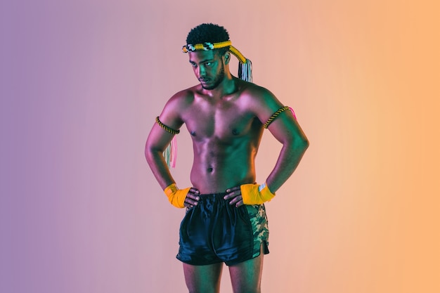 Muay thai. Young man exercising thai boxing on gradient wall in neon light. Fighter posing confident, training in martial arts in action, motion. Healthy lifestyle, sport, asian culture concept.