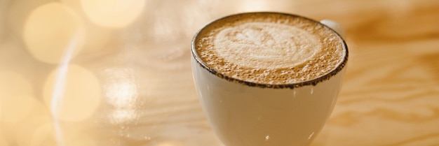 Mug of hot cappuccino on a wooden table in a cafe with yellow garland lights Web banner