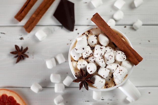 Mug of hot coffee with marshmallows sprinkled with chocolate, anise, cinnamon and a slice of dried grapefruit on white wooden table. Top view.