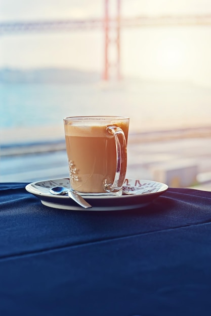 Mug of hot latte on table with blue tablecloth against of window with Lisbon bridge