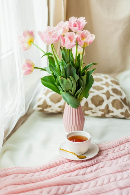 A mug of hot tea, a pink knitted blanket, a bouquet of tulips in a vase, a candle on the bed.