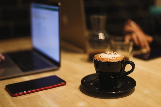 A mug of latte art coffee in shape of cute cat with laptop in the background.