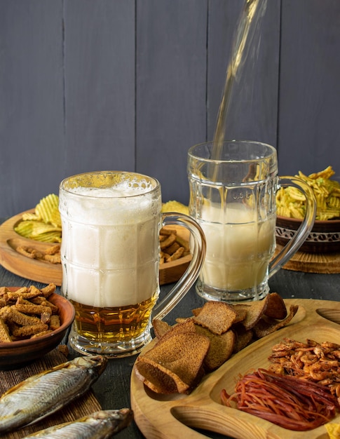 Mugs of cold beer with foam served on a wooden table with salty snacks in a wooden bowl