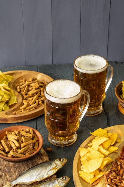 Mugs of cold beer with foam served on a wooden table with salty snacks in a wooden bowl