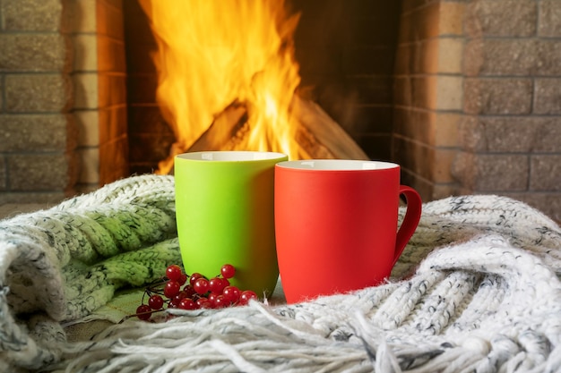 Mugs for tea or coffee and wool scarf before cozy fireplace